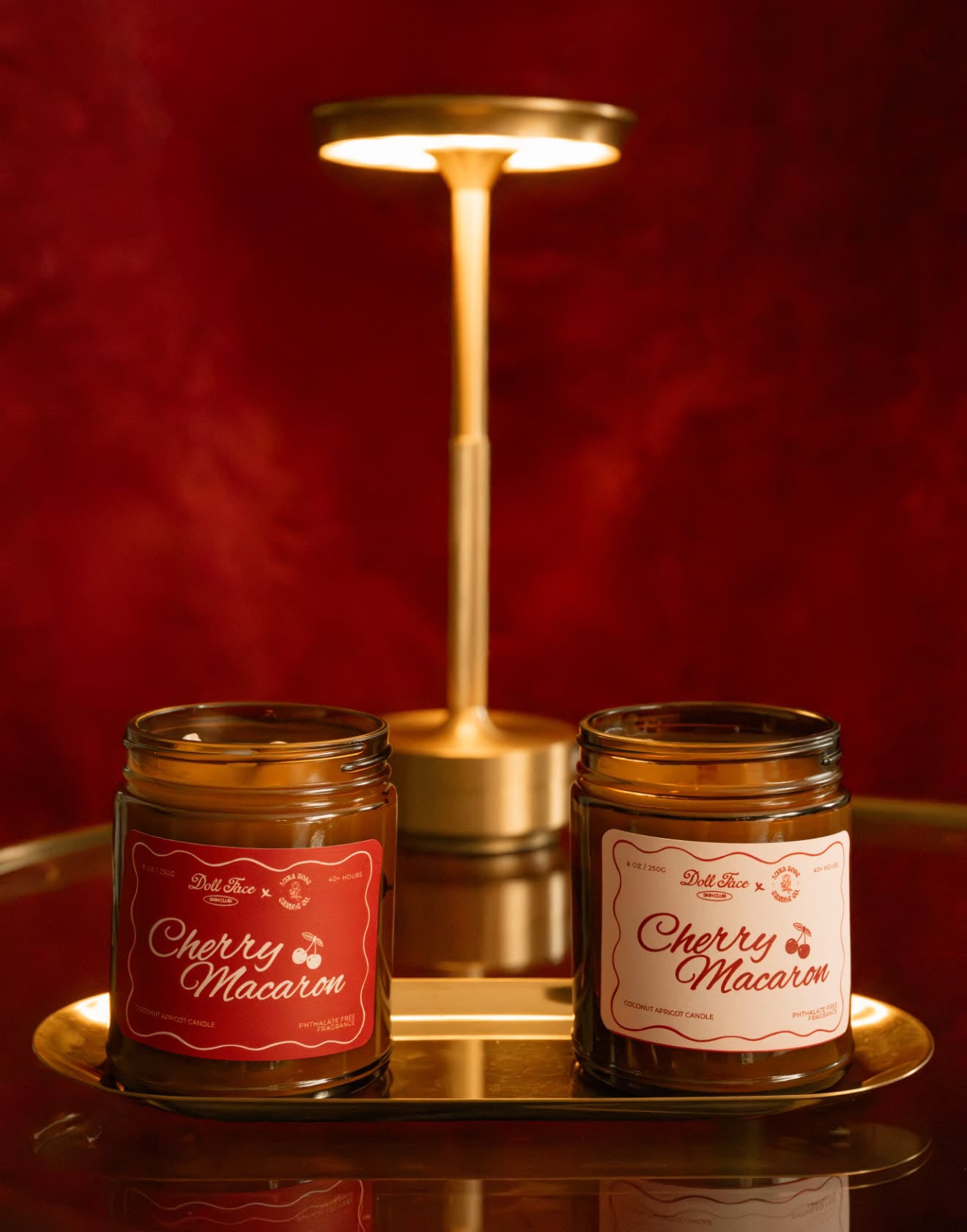Two amber glass jars of Cherry Macaron candles on a gold tray, with a matching gold table lamp behind them against a dark red textured background.