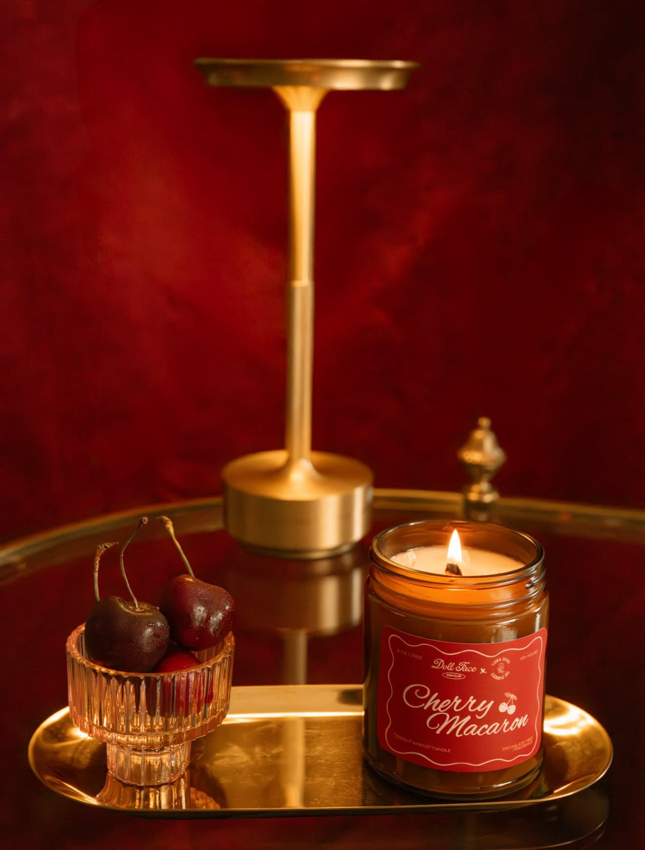A lit candle in a glass jar labeled 'Cherry Macaron' sits on a reflective gold tray beside a small glass bowl of cherries, with a gold candlestick holder and a small decorative object against a dark red background.