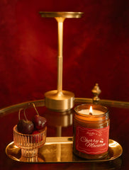 A lit candle in a glass jar labeled 'Cherry Macaron' sits on a reflective gold tray beside a small glass bowl of cherries, with a gold candlestick holder and a small decorative object against a dark red background.