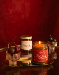 A lit jar candle labeled 'Cherry Macaron' sits on a reflective tray beside a closed matching jar and small glass bowls of cherries, against a deep red textured background.
