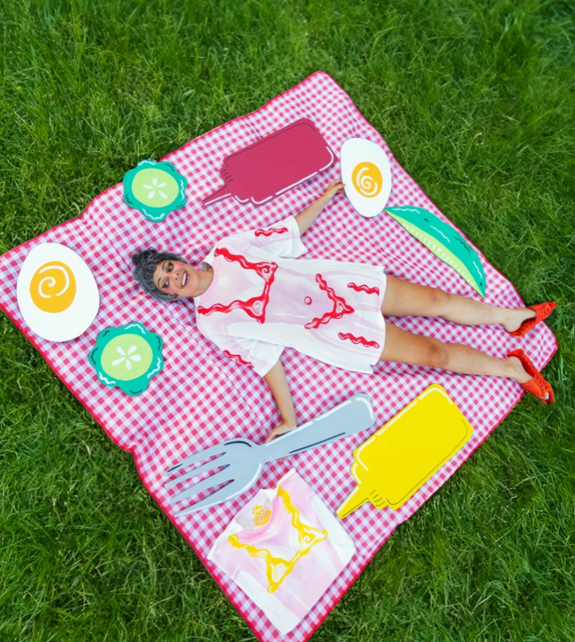 Person lying on a pink checkered picnic blanket with food-themed cutouts on grass