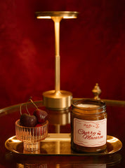 A candle in an amber glass jar labeled 'Cherry Macaron' sits on a reflective gold tray beside a small glass bowl of cherries, with a gold floor lamp and dark red velvet background.