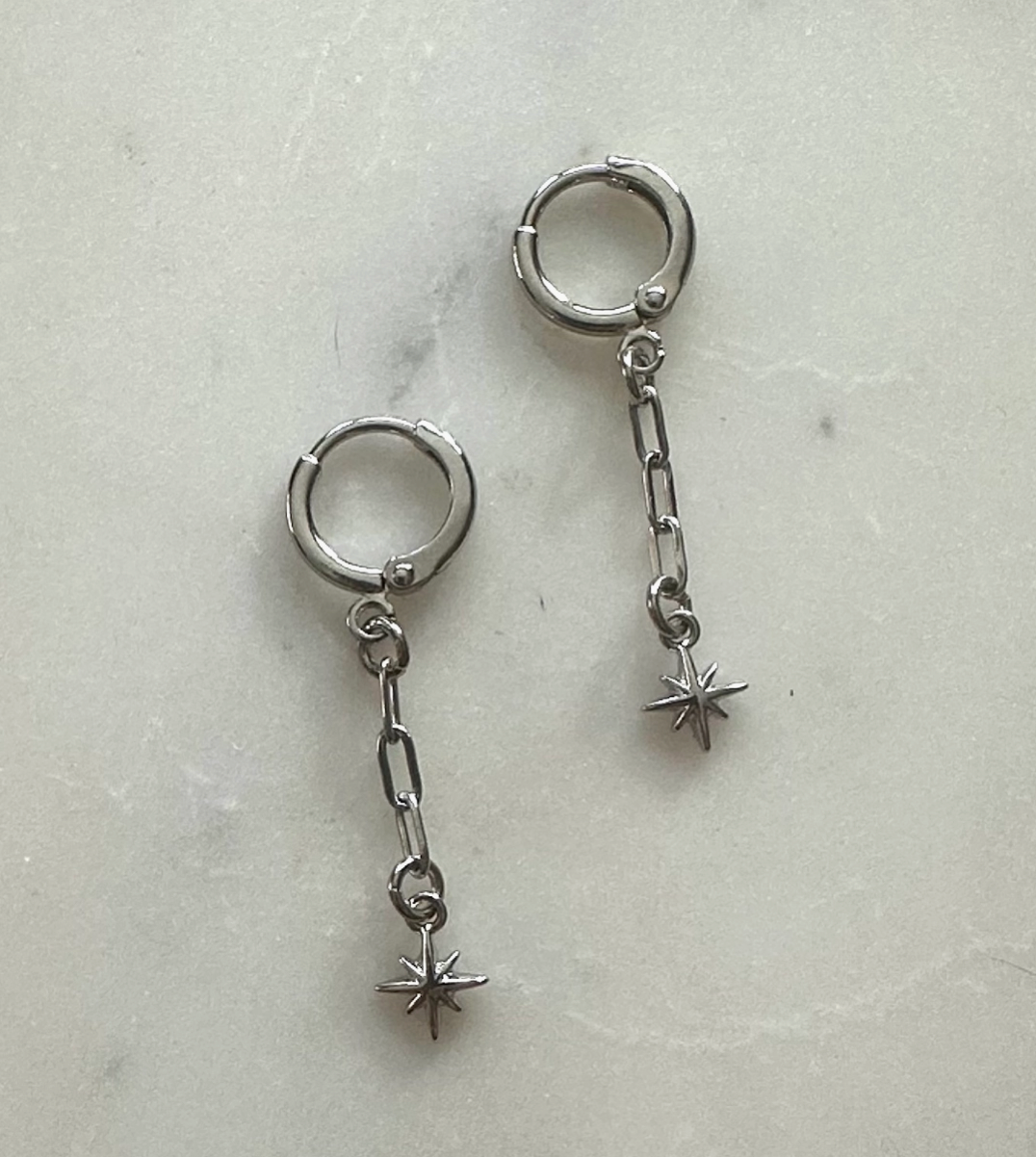 Pair of silver hoop earrings with chain and star-shaped charms on a light gray background